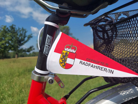 Ein Fahrradwimpel mit dem Schriftzug "Geprüfte/r Radfahrer/in" und dem Wappen des Landes Salzburg befestigt am Sitz eines Fahrrads.