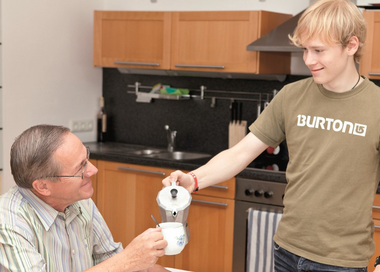 Pflegefit-Ausbildung. Ein junger Mann schenkt einem älteren Herrn Kaffee in seine Tasse. Am Tisch vor ihnen sind Kaffeeservice und Kuchen aufgebaut.