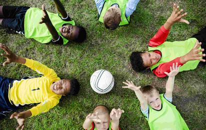 Sechs Buben liegen am Rücken in Gras und schupfen einander einen Fußball zu