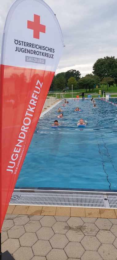 Schwimmer:innen mit Brett bei einer Schwimmübung. Links eine Beachflag des Salzburger Jugendrotkreuzes.