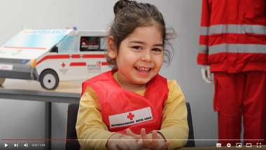 Ein Mädchen aus dem Kindergarten mit Rotkreuzweste erklärt wie man Hilfe holt