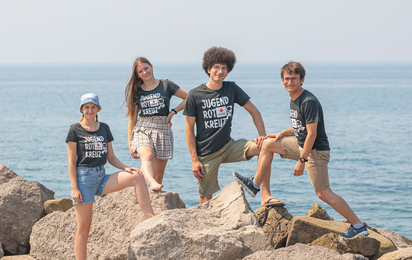 Zwei Campleiterinnen und zwei Campleiter stehen vor dem Meer im Jugendrotkreuz T-Shirt und lachen in die Kamera.