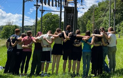 Viele Mädchen und Burschen stehen mit dem Rücker zum Betrachter Arm in Arm auf einer Wiese eines Hochseilgartens.