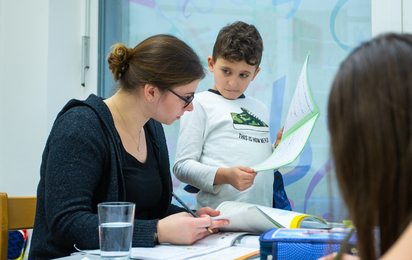 Lehrerin sitzt am Tisch und kleiner Junge steht neben ihr, welchem sie gerade etwas erklärt.