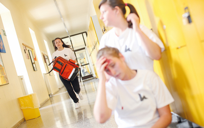 Eine Schüler*in ist verletzt und zwei andere Kinder leisten Erste Hilfe.